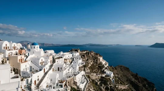 Whitewashed Cycladic village cascading down volcanic cliff overlooking azure Mediterranean waters